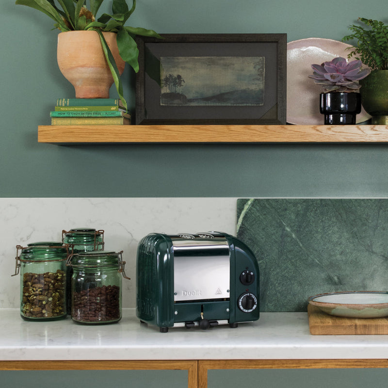 Dualit | 2 Slice Classic Toaster, Evergreen, on a marble counter with coffee jars, cutting boards, and a floating shelf adorned with decor items in a stylish kitchen setting.