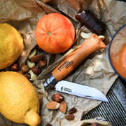 Opinel No 8 Carbon Steel Folding Knife with open blade, surrounded by lemons, nuts, and a chocolate bar, set on a wooden surface for a picnic.