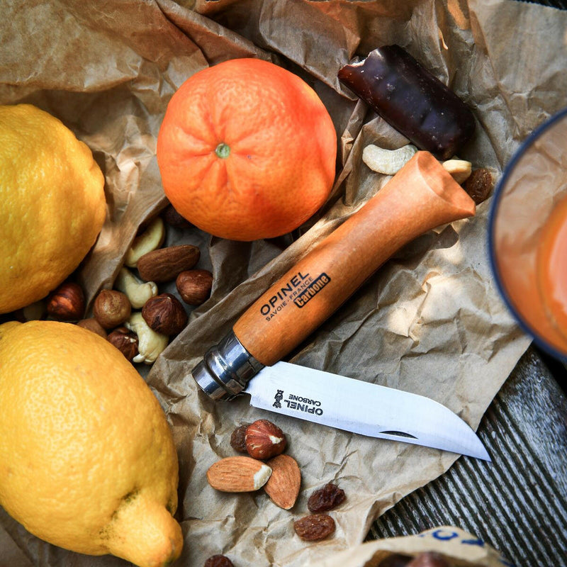 Opinel No 8 Carbon Steel Folding Knife with open blade, surrounded by lemons, nuts, and a chocolate bar, set on a wooden surface for a picnic.