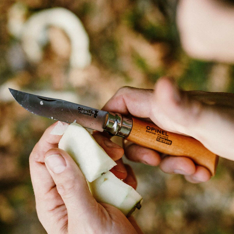 Hands using the Opinel No 8 Carbon Steel Folding Knife to carve a fruit, highlighting its wooden handle and robust design, ideal for culinary tasks.