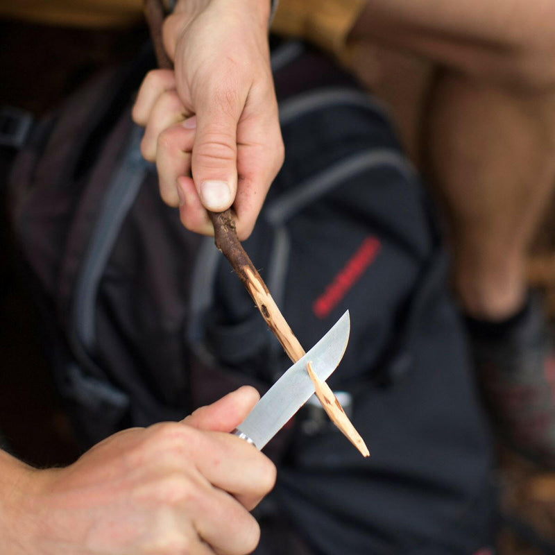 Person whittling wood with Opinel | No 8 Carbon Steel Folding Knife, suggesting outdoor use. Ideal for explorers and chefs; features Virobloc® safety ring.