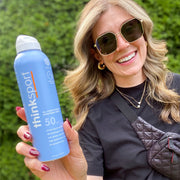 Woman smiling and holding Thinkbaby & Thinksport | All Sheer Mineral Sunscreen Spray SPF 50, emphasizing high-performance UVA/UVB protection, ideal for active, eco-conscious lifestyles.