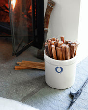 Farmhouse Pottery | Laurel Fireside Crock filled with kindling, featuring a blue wreath emblem, sits on a hearth with a fireplace and hearth tool nearby.