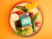 Spicewalla | Pescado Verde seasoning tin on a plate with green chiles, garlic, black peppercorns, shallots, cilantro, lime, and orange wedges.