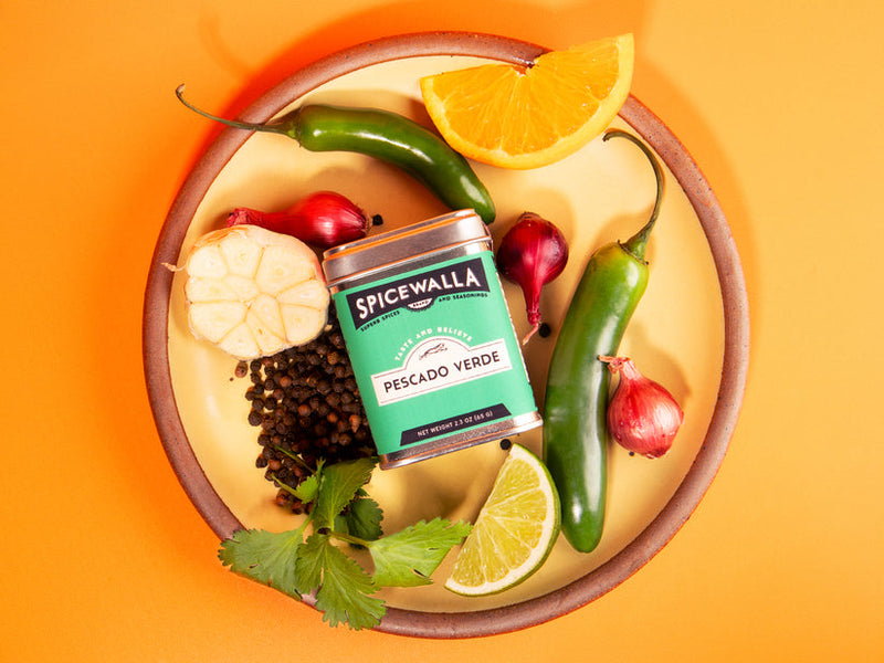 Spicewalla | Pescado Verde seasoning tin on a plate with green chiles, garlic, black peppercorns, shallots, cilantro, lime, and orange wedges.