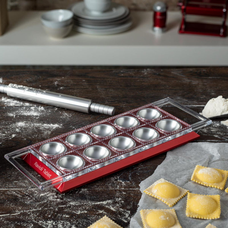 Marcato Ravioli Tablet Pasta Maker on a table, featuring a tray of ravioli, rolling pin, and tools for forming, cutting, and sealing ravioli.
