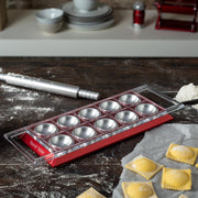 Marcato Ravioli Tablet - Powder Pink on a floured surface with ravioli mold, rolling pin, and freshly made ravioli, showcasing homemade pasta preparation.