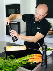 Man seasons casserole with Peugeot | Paris u'Select Pepper Mill, Satin Black, amidst fresh carrots and salad, showcasing fine-dining elegance in a modern kitchen.
