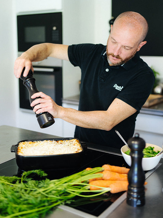 Man seasons casserole with Peugeot | Paris u'Select Pepper Mill, Satin Black, amidst fresh carrots and salad, showcasing fine-dining elegance in a modern kitchen.