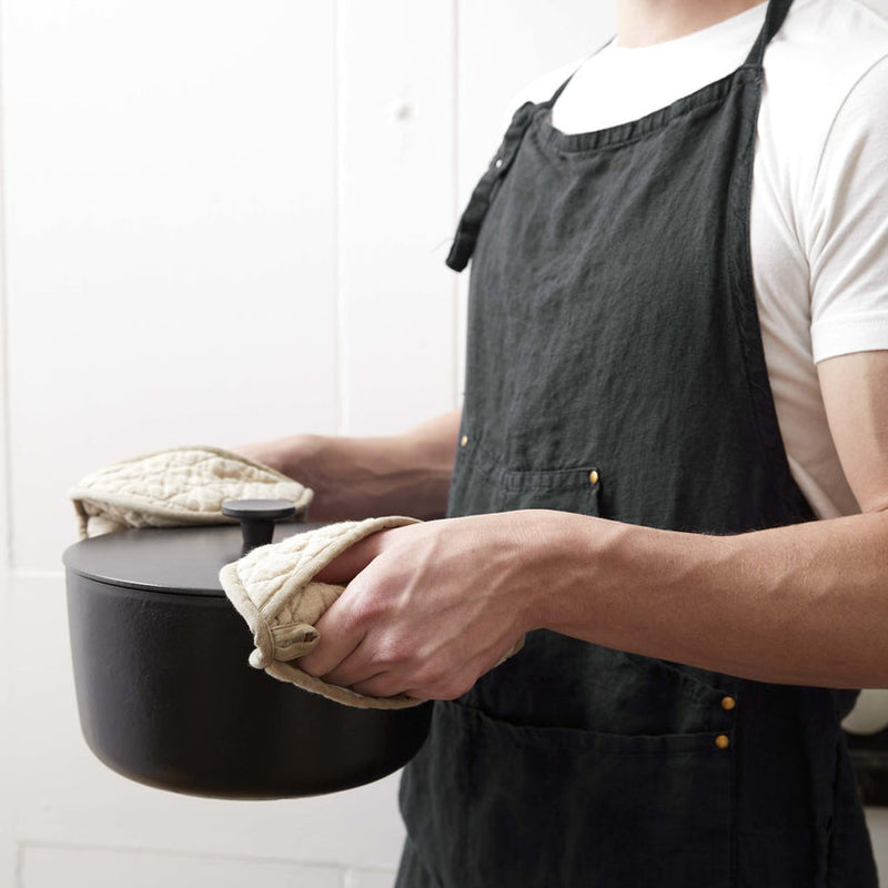 Person wearing the Sir|Madam Linen Grand Apron, holding a black Dutch oven with quilted mitts, showcasing adjustable neck loop and waist-level compartments.