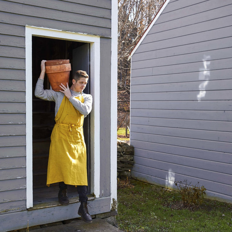 Gardener wearing the Sir|Madam Linen Grand Apron carries a terracotta pot, showcasing the apron’s practical pockets and adjustable neck, ideal for outdoor tasks.