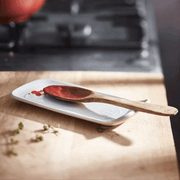 Emile Henry | Spoon Rest, Truffle holds a wooden spoon with tomato sauce on a kitchen countertop, complemented by scattered herb sprigs.