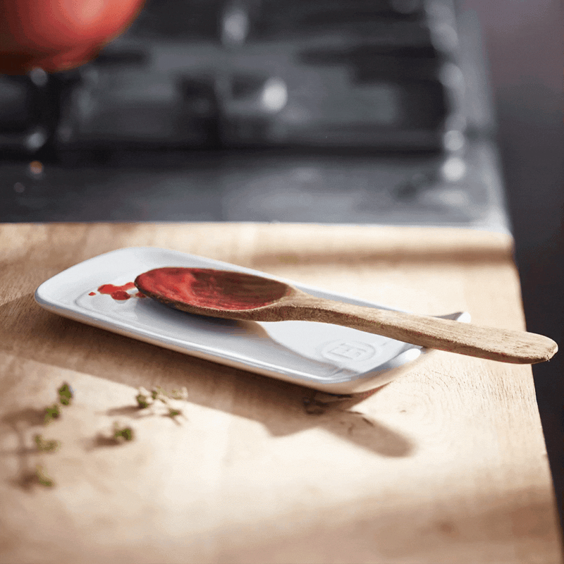 Emile Henry | Spoon Rest, Truffle holds a wooden spoon with tomato sauce on a kitchen countertop, complemented by scattered herb sprigs.