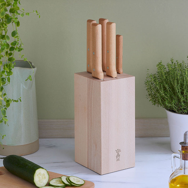 Opinel | 5-Slot Beech Wood Knife Block with light-wood handles, holding five knives, sits on a kitchen countertop beside a cucumber, herb plant, and olive oil.