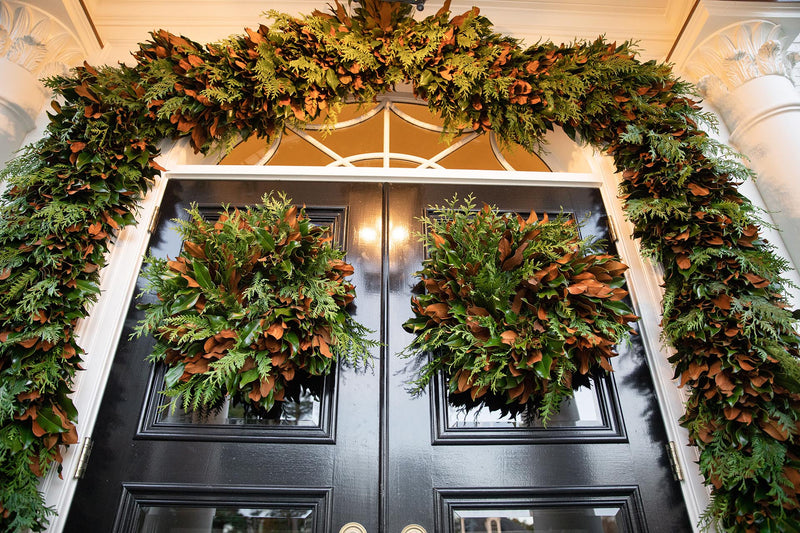 Conifer Magnolia Wreaths adorn glossy black doors with lush greenery, featuring cedar sprigs and magnolia leaves, creating an elegant, inviting entry.