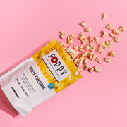 Poppy | Handcrafted Popcorns - White Cheddar spilling from a bag onto a pink surface, showcasing its crunchy, cheesy texture, ideal for snacking.