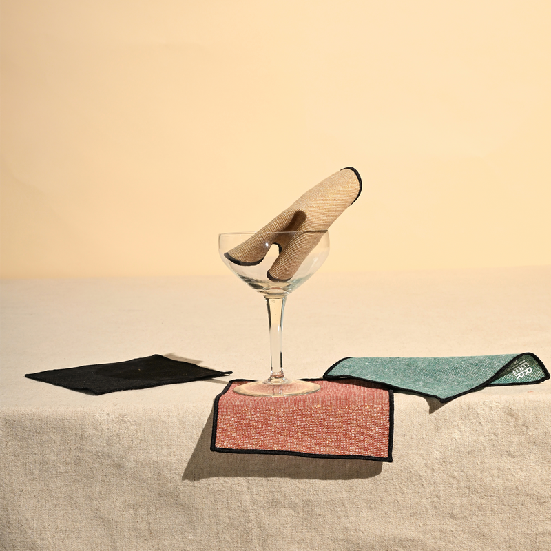 ATELIER SAUCIER - Luxxe Cocktail Napkins | Set of 4 displayed in a coupe glass, showcasing elegant, gold-spun fabric napkins bordered with black thread for a sophisticated touch.