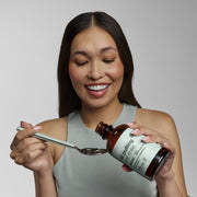 Young woman pouring Davids Hydroxi™ oil pulling oral rinse from an amber bottle into a spoon, designed for enhanced oral care with natural ingredients.