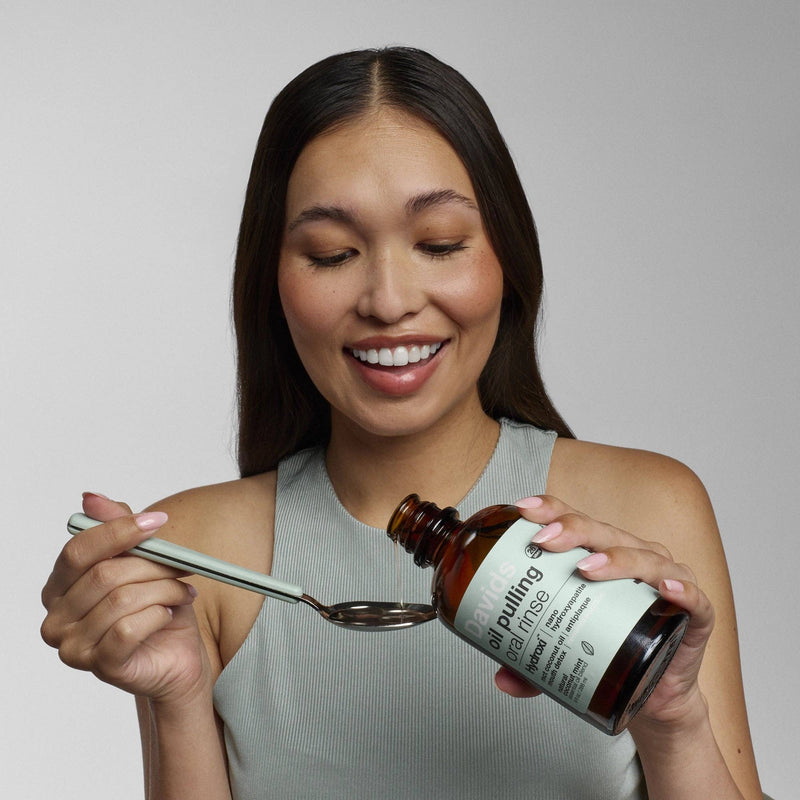 Young woman pouring Davids Hydroxi™ oil pulling oral rinse from an amber bottle into a spoon, designed for enhanced oral care with natural ingredients.