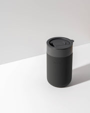 W&P - Ceramic Reusable Coffee Mug 16oz: Charcoal, featuring a silicone sleeve and lid, on a white surface. Ideal for eco-conscious, stylish coffee lovers.