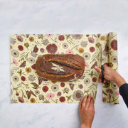 Bee's Wrap - Cut-to-Size Roll - Meadow Magic being trimmed to wrap a rustic dragonfly-stenciled bread loaf on a marble countertop, showcasing eco-friendly kitchen solutions.