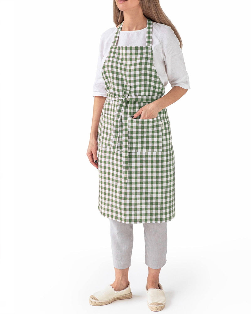 MagicLinen - Linen bib apron with pockets in Forest Green gingham, modeled by a woman. Features adjustable neck strap and large front pocket. Ideal for cooking in style.