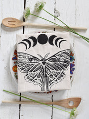Hearth and Harrow - Organic Cotton Luna Moth Tea Towel with moon phases, displayed on a floral plate, accompanied by wooden spoons and delicate white flowers.