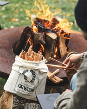 Farmhouse Pottery - Fatwood - 5lb. Bag with pine-resin sticks, ideal for starting fires, next to a burning fire pit. Hands reach for kindling sticks.