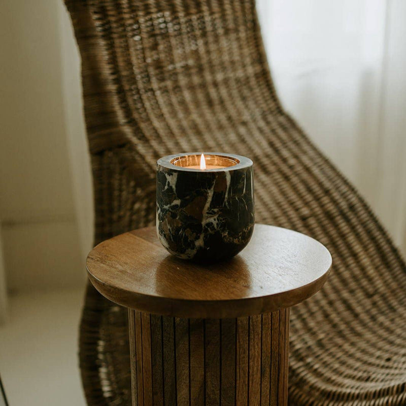 Last Candle Co - Portoro Marble Refillable Candle on a wooden table, featuring a dark stone vessel with gold veining, beside a wicker chair in a cozy interior.