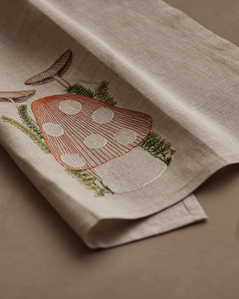 Coral & Tusk - Mushroom Tea Towel: Light-beige linen, embroidered with red toadstool, smaller mushrooms, green moss, ferns, and a ladybug, adding whimsy to your kitchen.
