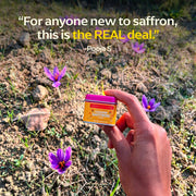 Hand holds a tin of Diaspora Spices Co. - Kashmiri Saffron (Grade A Threads) amid emerging saffron flowers, highlighting its single-origin authenticity and premium quality.