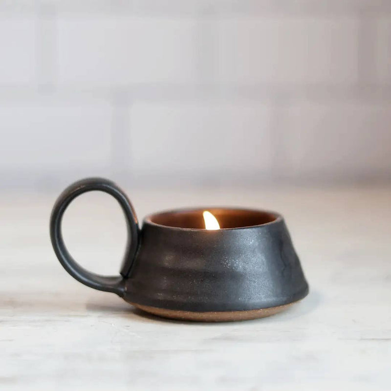 Gravesco Pottery - Tea Lite Candle Holder in Black, showcasing a lit candle inside the ceramic mug with a handle, set against a white-brick backdrop.