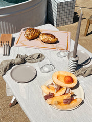 The MK Free Board™️, a plant-based cutting board, showcases grilled chicken, set amid a rustic outdoor table arrangement with cantaloupe, prosciutto, and elegant dining accessories.