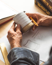 Person measuring a Farmhouse Pottery - Silo Pitchers: Petite ceramic cup at a design desk, with sketch, ruler, and books nearby, reflecting craftsmanship and design precision.