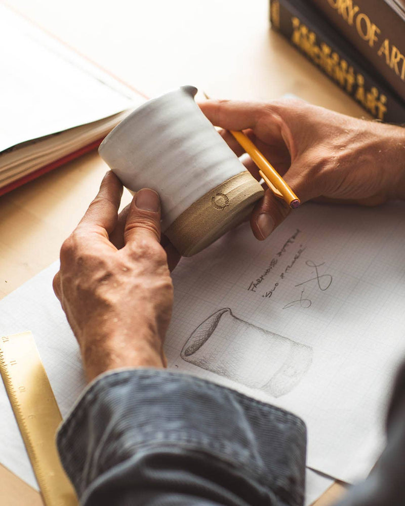 Person measuring a Farmhouse Pottery - Silo Pitchers: Petite ceramic cup at a design desk, with sketch, ruler, and books nearby, reflecting craftsmanship and design precision.
