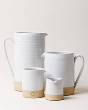 Farmhouse Pottery - Silo Pitchers: Petite. Four stoneware vessels with white glaze and exposed clay bases. Two pitchers behind, two creamers in front, featuring minimalistic design.