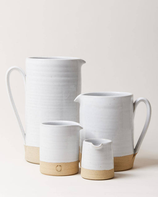 Farmhouse Pottery - Silo Pitchers: Petite. Four stoneware vessels with white glaze and exposed clay bases. Two pitchers behind, two creamers in front, featuring minimalistic design.