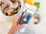 Person holding Mackenzie's Fisherman Hand Scrub tube, with shrimp, lemon, and basil in the background, suggesting its use for cleaning after handling seafood.