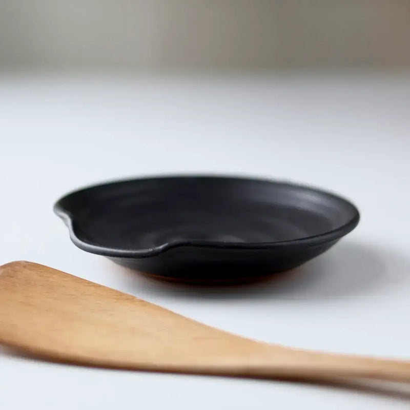 Gravesco Pottery - Handmade Pottery Spoon Rest in Black, a small, shallow dish with a spouted notch, sits beside a wooden spatula on a white surface.