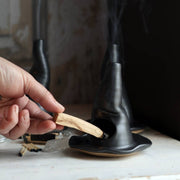 Gravesco Pottery - Witch Hat Incense Burner: A hand inserting palo santo into a black ceramic burner shaped like a melting candle, smoke rising, matches and quartz nearby.