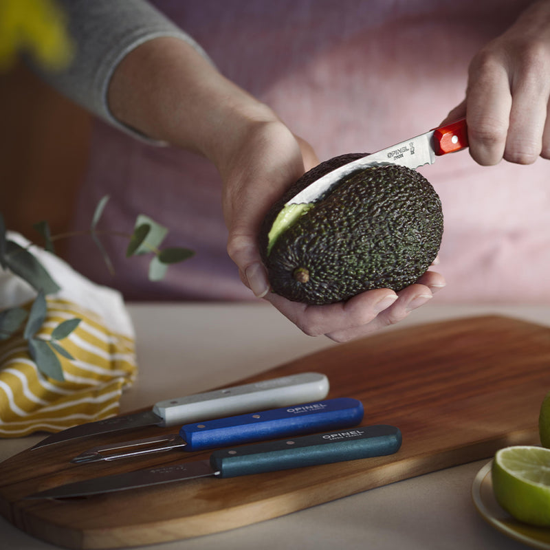 Close-up of Opinel Essential Small Kitchen Knife Sets Landscape, showcasing a person slicing an avocado with a red-handled knife, alongside three additional knives on a cutting board.