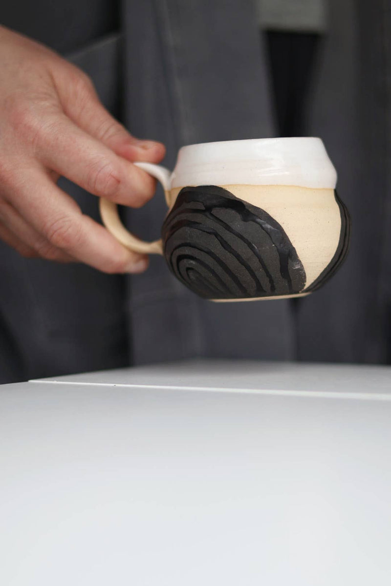 Close-up of a hand holding the Gravesco Pottery - Transformation Mug Petite, featuring a beige body with a black spiral motif, above a white tabletop.