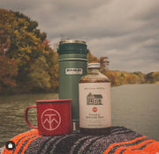 Pink House Alchemy - Pumpkin Butternut Spice Syrup, glass bottle on a blanket by water, alongside a travel mug and camp-style mug, in a fall-themed setup.