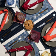 ATELIER SAUCIER - Clubhouse Cocktail Napkins: Set of 4, layered on a stylish table setting with patterned placemats, showcasing elegant tortoiseshell cutlery and vibrant coasters.