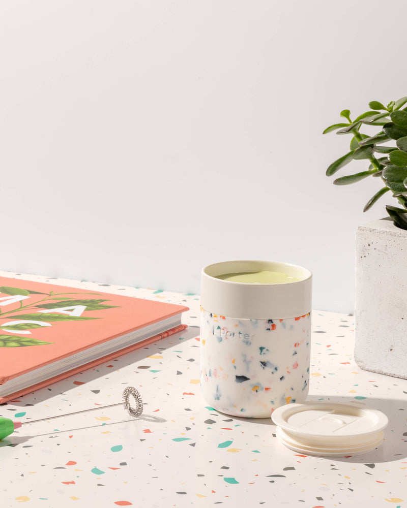 W&P - Reusable Ceramic Mug: Terrazzo Blush, 16 oz, filled with green liquid, beside a plant, book, and notebook, highlighting its eco-friendly and durable design.