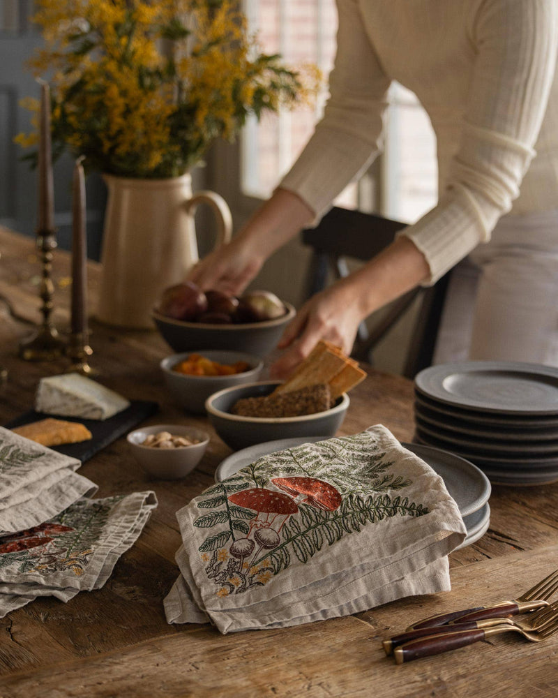 Mushrooms and Ferns Bouquet Dinner Napkin on rustic table with stoneware, cheese, and flowers. Cream-sweatered individual arranges bowls, enhancing a cozy, natural setting.