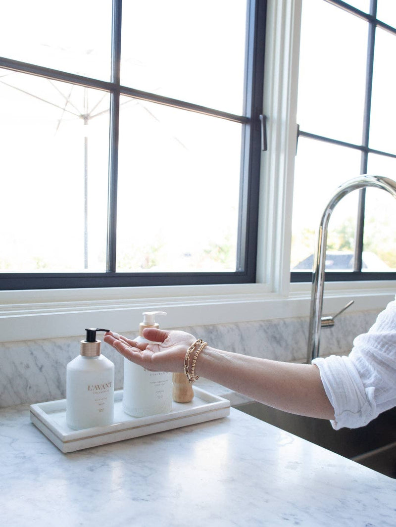 Person applying L'AVANT Collective - Fresh Linen Hand Lotion from a white bottle with gold text, near a sink on a white marble countertop.