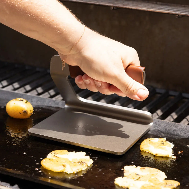 A hand uses the Made In | Grill Press with a leather handle to flatten potato slices on a hot griddle, highlighting its sturdy, flat-bottomed design.