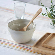 bambu® - Bamboo Serving Spoon rests in a ceramic bowl with a wooden spoon, surrounded by bamboo trays, a ribbed glass, and a vase with greenery.