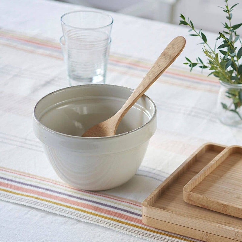 bambu® - Bamboo Serving Spoon rests in a ceramic bowl with a wooden spoon, surrounded by bamboo trays, a ribbed glass, and a vase with greenery.